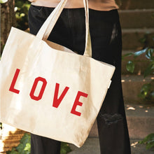 Load image into Gallery viewer, Natural cotton canvas tote bag with red "LOVE" lettering and shoulder straps, shown worn over the shoulder. 