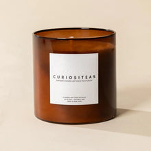 Load image into Gallery viewer, Poured NY Curiositeas Candle | Two Styles, white wax in a cylindrical translucent dark amber-brown glass vessel with a square white label with "CURIOSITEAS" in dark lettering displayed against a cream background. 
