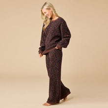 Load image into Gallery viewer, Person smiling and walking barefoot in an espresso-colored lounge set featuring a crewneck sweater and matching wide-leg pants with a black leopard print pattern. The outfit has ribbed cuffs and hems, shown against a soft beige background. 