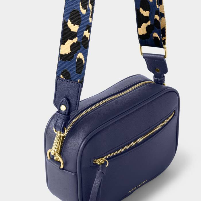 A navy-colored crossbody bag featuring  gold-tone hardware, and a blue leopard print strap, on a white background.