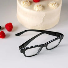 Load image into Gallery viewer, Black chunky square reading glasses with black and white checkered pattern along top edge, displayed on white surface with a cake and raspberries in the background.