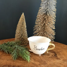 Load image into Gallery viewer, A white teacup ornament with a gold glitter handle, a clear footed bottom, and &quot;Bourbon&quot; in black text. On a wooden surface with a green fir tree cutting , and two decorative frosted Christmas trees. Against a dark background.  