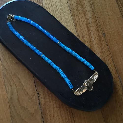 A necklace with a gold-tone beetle pendant and blue beads displayed on a black display on a wooden surface.