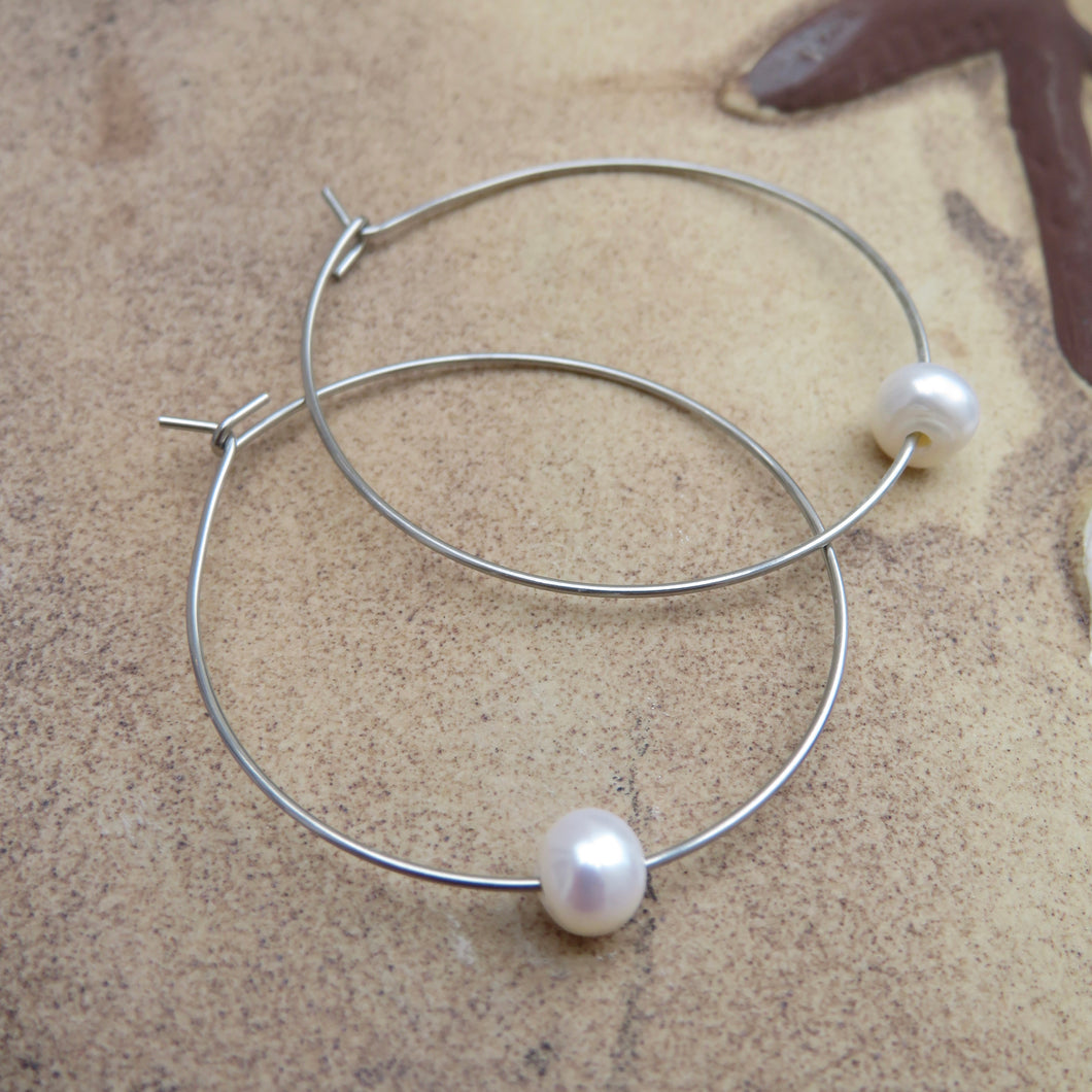 Silver hoop earrings with a single pearl on a textured beach surface