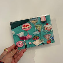 Load image into Gallery viewer, Art print featuring illustrated cocktail glasses, olives, makeup items, and desserts on a teal background, shown held in hand against a white background.