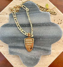 Load image into Gallery viewer, Back side of gold shield-shaped pendant on a gold paper clip chain necklace, featuring engraved text on the pendant, displayed on a quatrefoil-shaped gray stone tile layered over a beige travertine slab on a wooden surface.  
