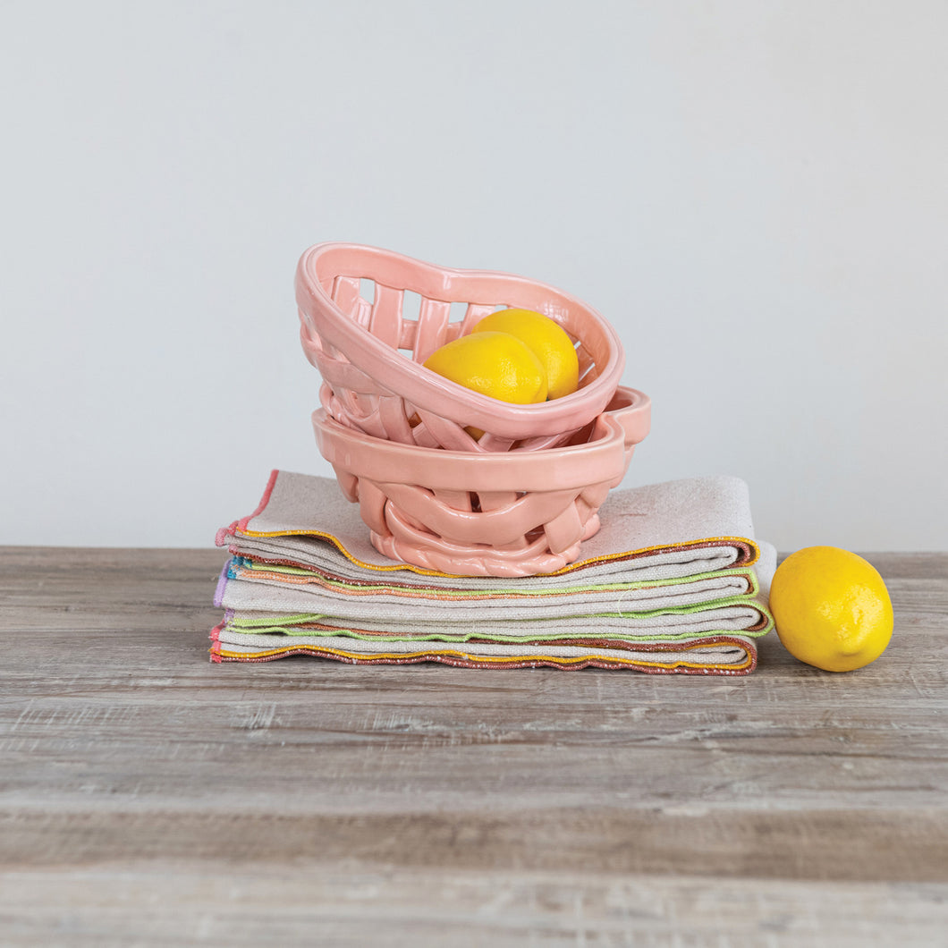 Stacked  pink heart-shaped stoneware fruit basket  with a woven design and two lemons inside , displayed on a stack of beige napkins with colorful trim. Lemon resting beside stack go napkins. On a wooden surface, with a white background behind. 