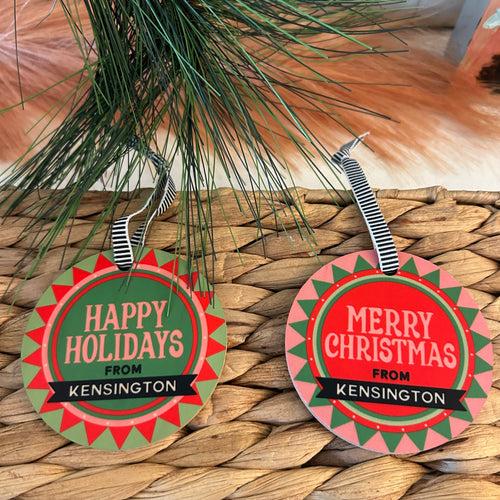 Two circular ornaments sitting on a brown reed woven basket surrounded by pine needles. One ornament is green and reads 