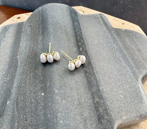 Pair of gold post earrings each featuring three small white pearls arranged in a row, displayed on a quatrefoil-shaped gray stone tile layered over a beige travertine slab.