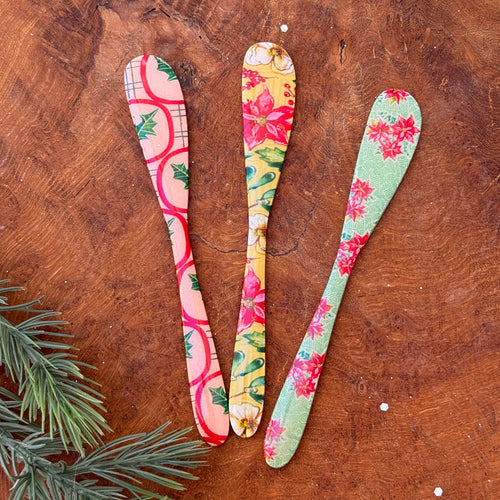 A pink canapé knife with a red and green mistletoe design. A yellow canapé knife with a pink, white, and green floral design.
A teal canapé knife with a pink floral design. On a brown wooden surface with green fir tree cuttings.