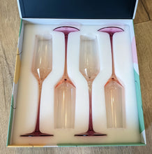 Load image into Gallery viewer, Set of four hand-blown crystal champagne flutes with pink gradient stems and bases, displayed in a fitted white foam insert inside a decorative gift box on a wooden surface.