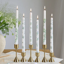 Load image into Gallery viewer, Six lit white taper candles with assorted floral and botanical designs, displayed in gold candle holders on scattered paper pages. 