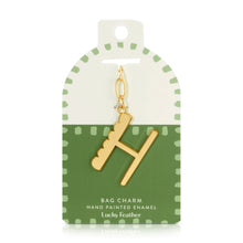 Load image into Gallery viewer, 14k gold-plated hand-painted enamel initial bag charm featuring the letter H with a scalloped edge design and a brushed gold finish, attached to a key ring and clasp. Displayed on a white and green Lucky Feather card with light green dotted brush-stroke borders labeled &quot;Bag Charm Hand Painted Enamel.&quot; Displayed on a white background. 