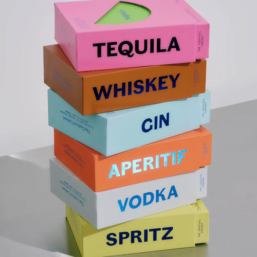 Stack of colorful cocktail recipe card boxes labeled Tequila, Whiskey, Gin, Aperitif, Vodka, and Spritz. Each box features bold black or blue lettering and is displayed against a white background. 