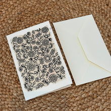 Load image into Gallery viewer, Colby Cards, lifestyle image of a white Lokta paper greeting card featuring black floral and leafy vine outlines on a cream panel with softly textured, uneven edges, displayed beside a white envelope on a brown woven mat.