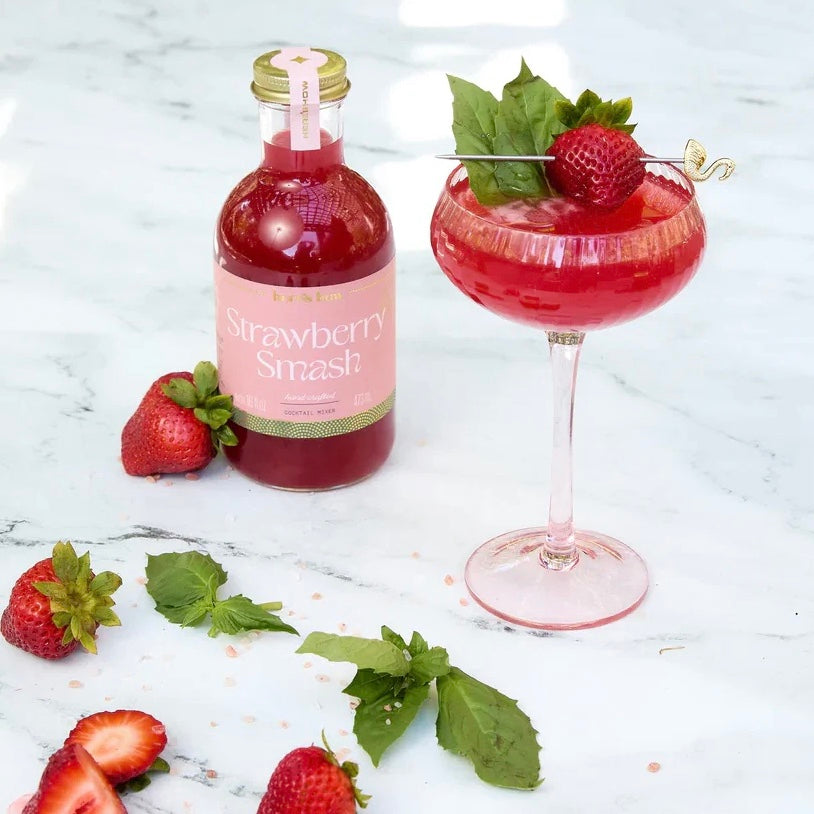 Bottle and glass of red  strawberry smash cocktail mixer with fresh strawberries and mint leaves on a marble surface.