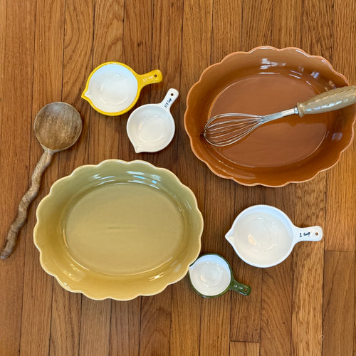 Olive-colored and terracotta-colored scalloped stoneware dishes displayed on a wooden surface with a whisk, wooden spoon, and ceramic measuring cups in white, yellow, and green.
