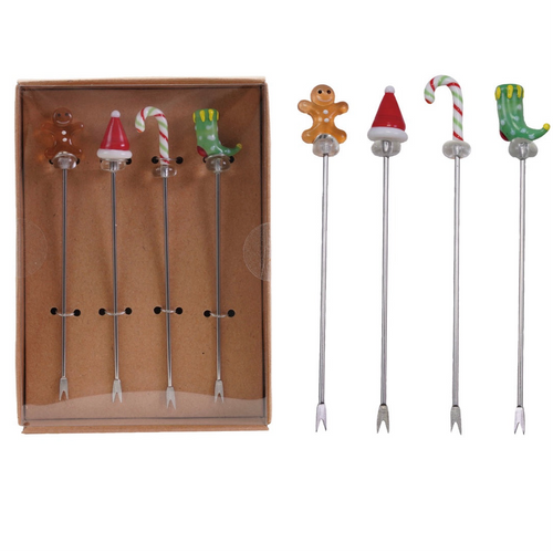 Two sets of four holiday glass cocktail picks with blown-glass toppers including gingerbread men, Santa hats, candy canes, and green elf boots, displayed in a brown box and arranged on a white background.  