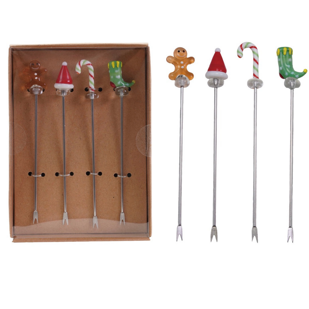 Two sets of four holiday glass cocktail picks with blown-glass toppers including gingerbread men, Santa hats, candy canes, and green elf boots, displayed in a brown box and arranged on a white background.  