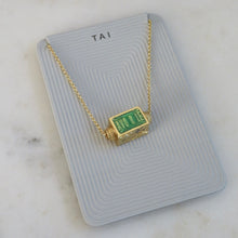 Load image into Gallery viewer, Gold-plated rectangular spinner pendant necklace with green enamel and raised lettering, displayed on a light gray TAI jewelry card on a white marble background.  