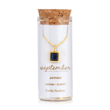 Load image into Gallery viewer, September birthstone necklace featuring a square dark blue sapphire glass crystal pendant framed in 14K gold-dipped metal, displayed inside a clear glass bottle with a cork lid and a label card reading "September - Sapphire - Wisdom + Clarity," displayed against a white background. 