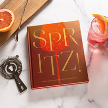 Load image into Gallery viewer, "Spritz!" drink recipe book with orange cover styled beside cocktail and bar accessories.
