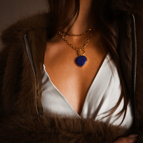 Person wearing a fur coat and white blouse with a gold chain necklace featuring a blue lapis stone pendant.