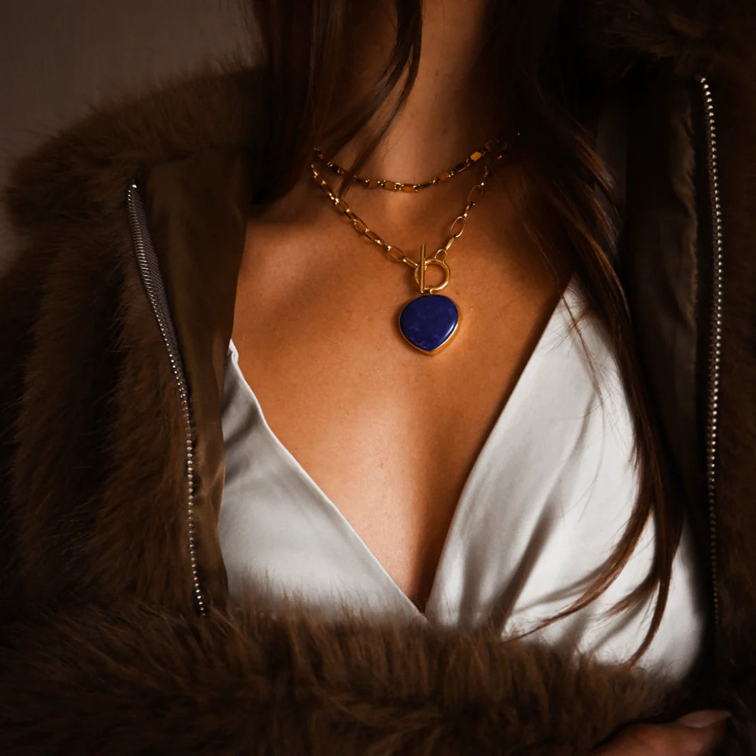 Person wearing a fur coat and white blouse with a gold chain necklace featuring a blue lapis stone pendant.