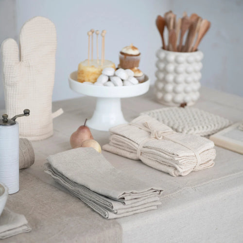 A set of beige linen and cotton cocktail napkins displayed on a table with other kitchen items in the background.