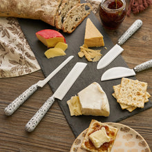 Load image into Gallery viewer, Four piece black-and-white tile-patterned stainless steel cheese knife set arranged on a slate board with assorted cheeses, crackers, bread, and a jar of preserves, displayed on a wooden surface.  