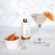 Load image into Gallery viewer, White cotton cocktail napkins with a gold border and gold outlined stack-of-glasses illustration set on a gold holder, displayed on a white marble surface next to a martini with an orange slice garnish and a white bowl of orange slices, both resting on matching white cocktail napkins, against a white tile background. 