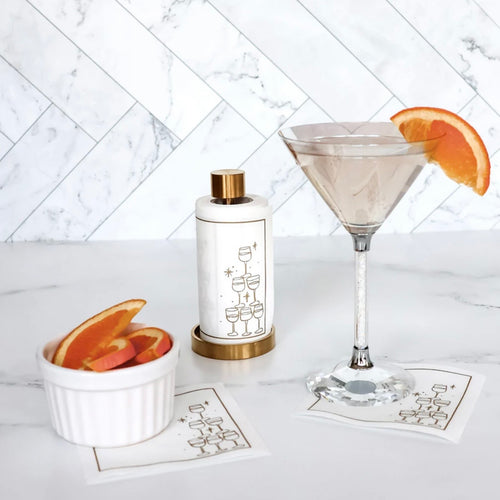 White cotton cocktail napkins with a gold border and gold outlined stack-of-glasses illustration set on a gold holder, displayed on a white marble surface next to a martini with an orange slice garnish and a white bowl of orange slices, both resting on matching white cocktail napkins, against a white tile background. 