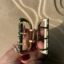Load image into Gallery viewer, Leopard-print half-moon metal claw clip with gold hardware held in a hand against a beige textured surface with soft curved light reflections. 