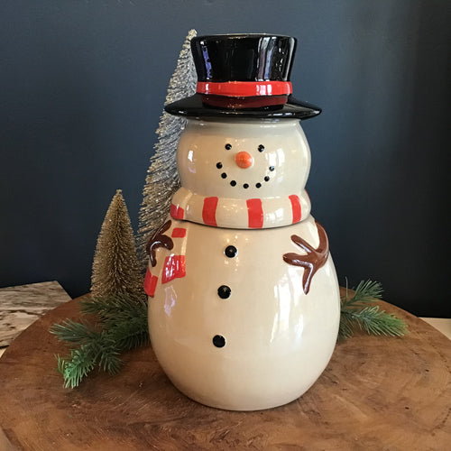White snowman cookie jar with a black top hat with a red stripe, a red and white striped scarf, an orange carrot nose, brown tree- like arms , and black dots for the eyes, smile, and buttons. On a wooden surface with frosted decorative Christmas trees and green fir tree cuttings. Against a blue wall. 