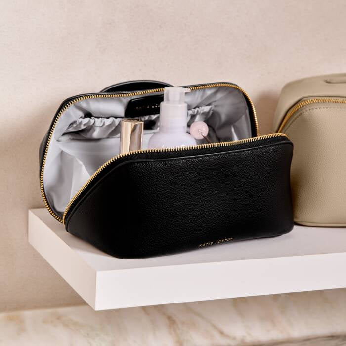 An opened black makeup bag with a gold zipper. Toiletries inside. A cream colored makeup bag with a gold zipper beside the black makeup bag.  Both sitting on a white floating shelf , with a cream color wall behind.