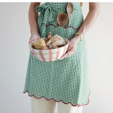 Load image into Gallery viewer, Holiday Apron, lifestyle photo showing the cropped view of a model wearing a green cotton gingham waist apron with red scalloped trim, holding a pink-and-white striped bowl filled with baked goods, with two wooden spoons tucked into the red-trimmed bib pocket, against a cream background. 
