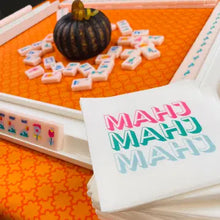 Load image into Gallery viewer, White cocktail napkins printed with "MAHJ" in pink, green, and blue, displayed beside white Mahjong racks with tiles and scattered pieces surrounding a small decorative pumpkin on an orange patterned surface. 