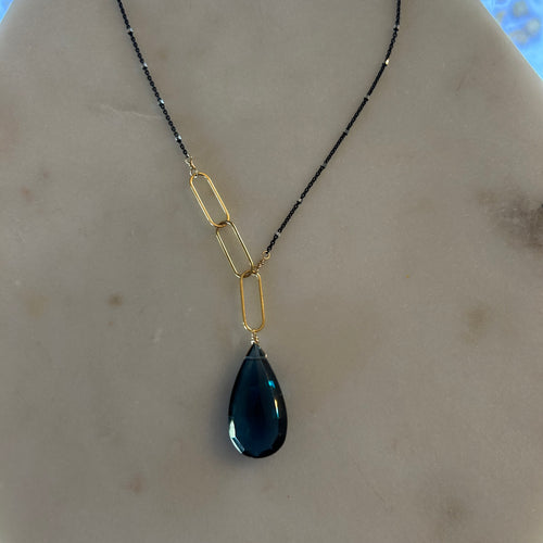 Black chain necklace with an asymmetrical paperclip gold chain one one side that drops down to a large blue teardrop gem. 