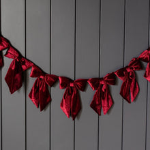 Load image into Gallery viewer, Velvet Bow Garland, a row of matching deep red velvet bows with long hanging tails, hanging in a gentle arc against a gray panel wall. 