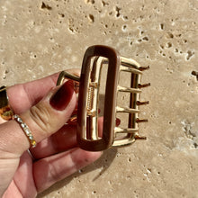 Load image into Gallery viewer, Tan rectangular metal claw clip with gold hardware held in a hand against a beige travertine surface with soft shadows. 