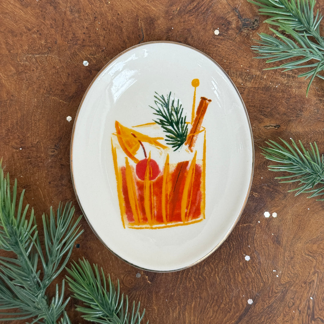 White, oval, ceramic dish with an orange colored glass with an orange drink design. A red cherry, an orange slice, a brown cinnamons stick, an orange stir pick, and a green fir garnish inside glass. On a wooden surface with fir tree cuttings. 