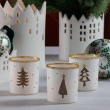 Load image into Gallery viewer, Three white tea light holders with gold glitter rims and simple black Christmas tree designs, styled with white ceramic village houses and decorative ornaments on a light surface. 