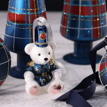 Load image into Gallery viewer, Teddy Bear Glass Ornament, lifestyle image of a seated white bear glass ornament in a metallic blue uniform and tall hat with gold detailing and round red jewel accent, displayed at an angle on a light tabletop among red and blue plaid cone tree decorations, matching ornament balls, and navy ribbon.