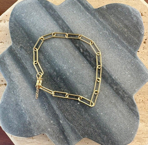 Gold-finish elongated paperclip chain bracelet with an extender, displayed on a overlapping gray quatrefoil stone tile and a beige travertine slab.