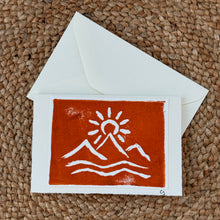 Load image into Gallery viewer, Colby Cards, lifestyle image of a white Lokta paper greeting card featuring an red-orange rectangular print with white mountain peaks, a sun, and wavy lines, displayed horizontally with a white envelope on a brown woven mat.