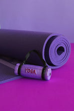 Load image into Gallery viewer, Yoga Mat Ornament, lifestyle image of a miniature rolled purple glass yoga mat with a textured surface, black carrying straps, and glittery red “YOGA” lettering, displayed beside a full-size rolled purple mat on a bright magenta surface against a purple background.