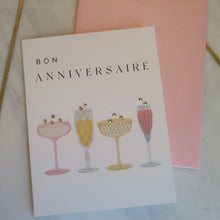 Load image into Gallery viewer, Greeting card with &quot;Bon Anniversaire&quot; text and illustrations of champagne glasses with sparkly bubbles on a pink envelope and marble background.