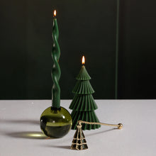Load image into Gallery viewer, Wave tree candle snuffer in a gold finish designed with a wave-inspired pine tree shape, displayed on a white surface beside two lit green candles-one in a round green glass holder with a twisted taper and one shaped like a pine tree- against a dark background. 
