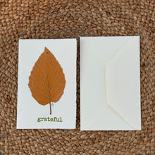 Load image into Gallery viewer, Colby Cards, lifestyle image of a white Lokta paper greeting card featuring a real pressed golden-orange leaf above the green word “grateful,” displayed beside a white envelope on a brown woven mat.
