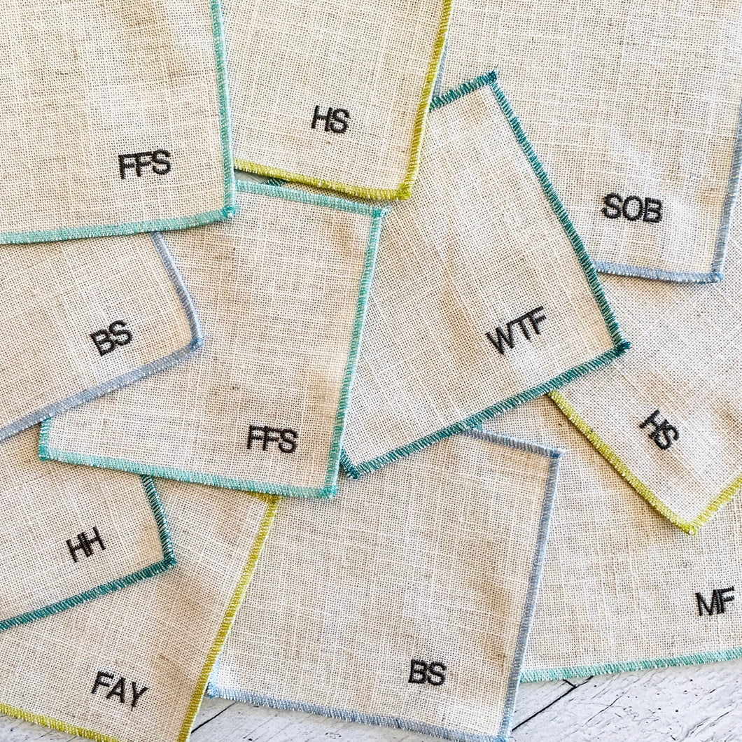 Collection of biege linen coasters with embroidered text and initials on a light background